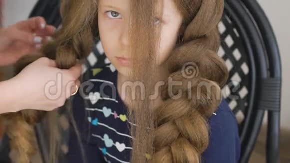 不开心的小女孩妈妈或理发师给她的辫子编辫子棕色头发视频的预览图
