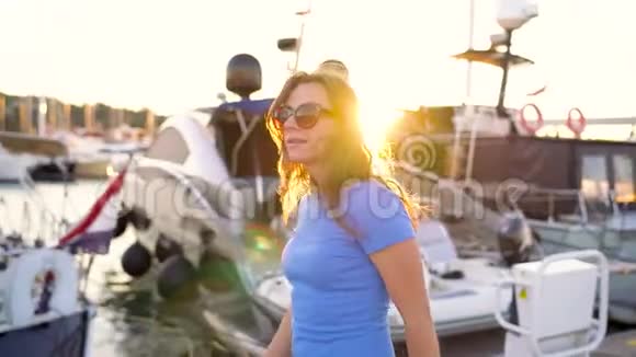 戴墨镜的女人在日落时带着很多游艇和船沿着码头走关门慢动作视频的预览图