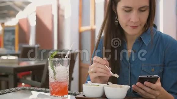 布鲁内特女孩坐在外面的咖啡馆里吃冰淇淋时使用智能手机4k视频的预览图
