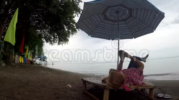 成熟的女人在热带海滩上躺在遮阳伞下用移动智能手机浏览互联网视频的预览图