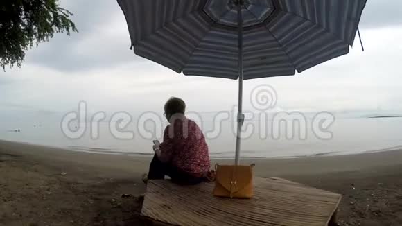 成熟女性盘腿坐在遮阳伞下用手机上网视频的预览图