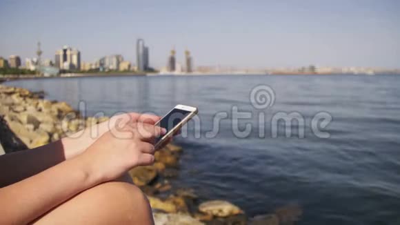 年轻女性使用智能手机坐在海边的堤岸和梅格波利斯摩天大楼上视频的预览图