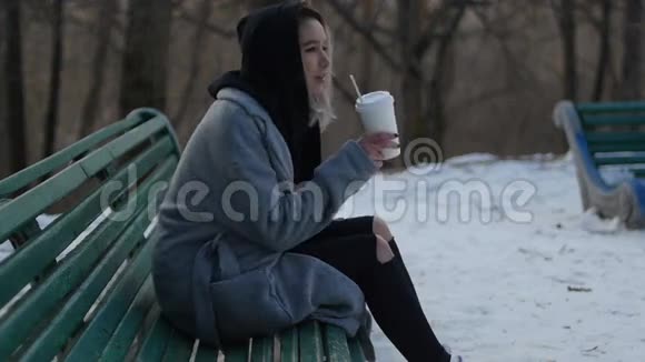 坐在长凳上的一个冬天公园里喝咖啡的小女孩视频的预览图