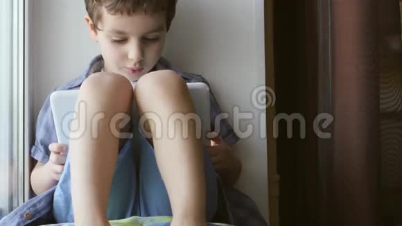 可爱的小男孩在家的窗台上使用白色平板电脑视频的预览图
