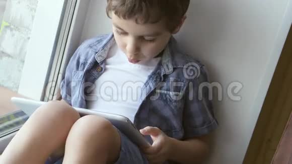 波特瑞特可爱的小男孩在家的窗台上使用白色平板电脑视频的预览图