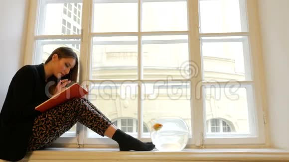 美丽梦幻的年轻女士写在笔记本上在窗台上摆姿势上面放着金色的鱼视频的预览图