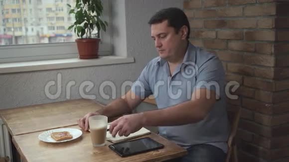 早上在酒店喝咖啡吃烤面包的成年男性每天都在桌子和平板上视频的预览图