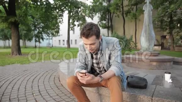 年轻人穿着牛仔裤夹克坐在花岗岩圆面上打电话自拍视频的预览图