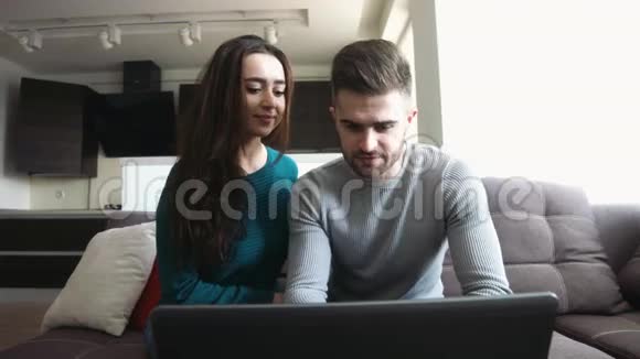 一对漂亮的夫妇坐在沙发上年轻人用笔记本电脑查看他的电子邮件和女朋友聊天亲吻她视频的预览图