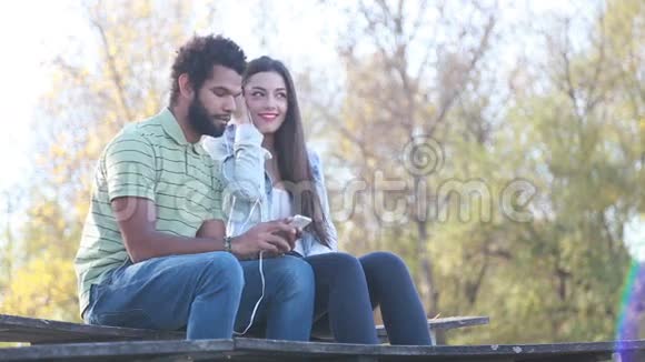 可爱的夫妇坐在长凳上用共用耳机听音乐视频的预览图