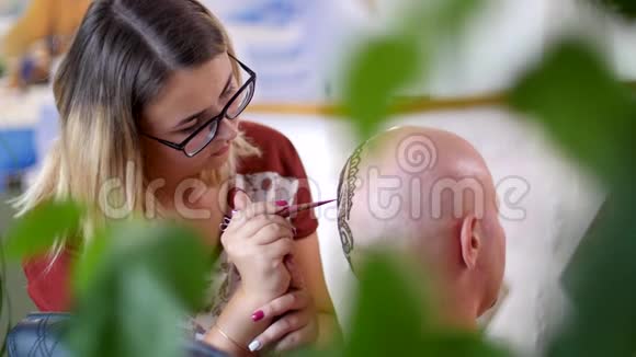 一个女孩纹身大师迈亨迪艺术家在秃顶白种人的头皮上画指甲花纹身穿着一件蓝色衬衫视频的预览图