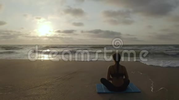 美丽的年轻女子在海滩上做瑜伽同时观看日落视频的预览图
