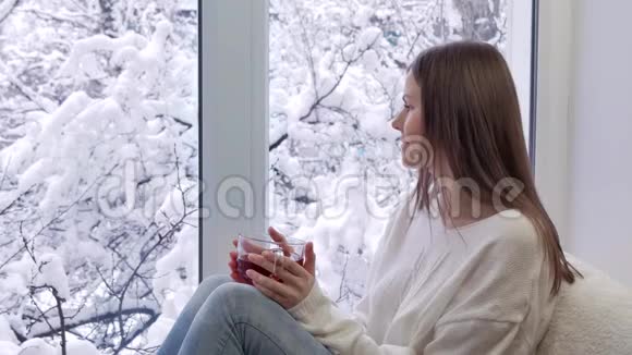 美丽的女孩坐在窗台上喝着茶看着窗户冬天在外面视频的预览图