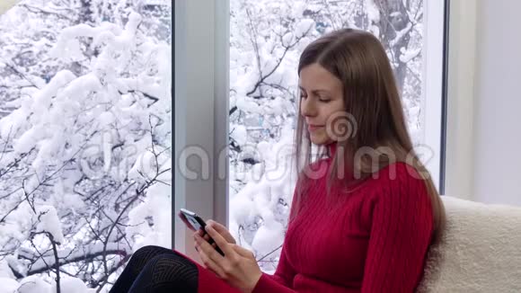 穿红色裙子的漂亮女孩坐在窗台上用智能手机冬天在外面视频的预览图