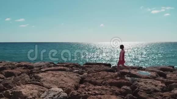 穿着红色衣服的女人走在岩石码头海滩上海浪拍打着悬崖阳光明媚的日子里阳光明媚太阳之路蓝色的倒影视频的预览图