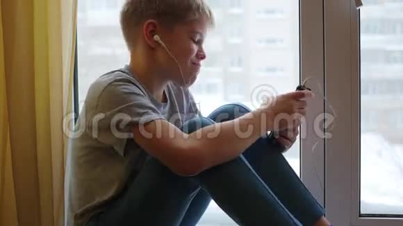 孩子坐在窗台上用耳机听智能手机上的音乐视频的预览图