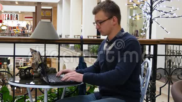 戴眼镜的年轻人坐在商场的桌子旁用笔记本电脑工作视频的预览图