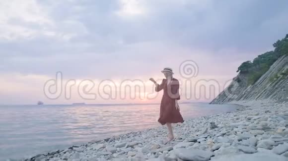 一个穿着红色夏装戴着草帽的漂亮姑娘小心翼翼地爬在岩石海岸的石头上视频的预览图