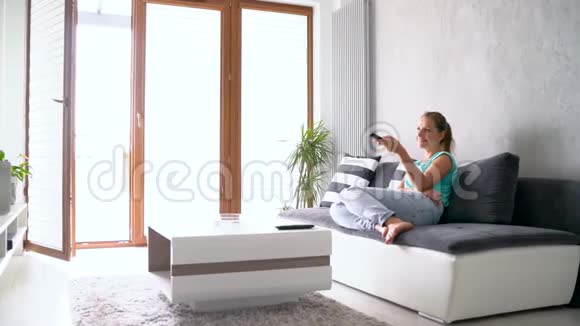 年轻漂亮的女人躺在沙发上看电视她无限期地打开遥控器追踪镜头视频的预览图