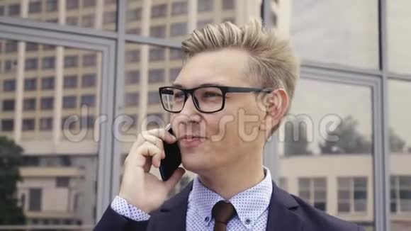 年轻英俊的高加索商人戴着眼镜在办公楼前用手机说话男性行政主管视频的预览图