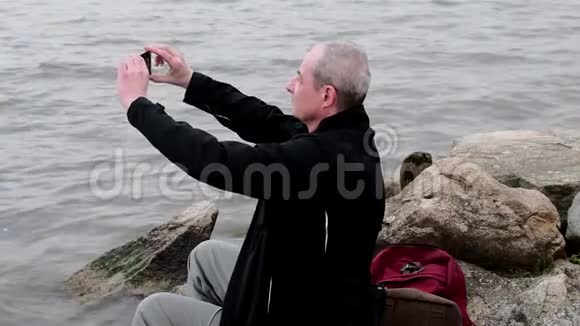 坐在湖岸上的中年老人孤独成熟的男人在银行拍自拍照片概念视频的预览图