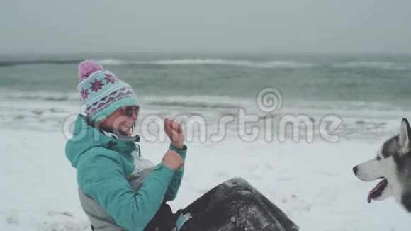 在暴风雪中年轻的女性和西伯利亚哈士奇狗在海滩上玩耍和奔跑动作缓慢视频的预览图