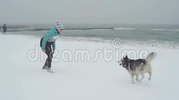 在暴风雪中年轻的女性和西伯利亚哈士奇狗在海滩上玩耍和奔跑动作缓慢视频的预览图