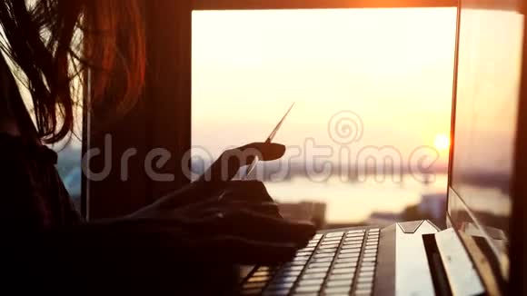 在美丽的日落和令人惊叹的阳光下女孩在城市背景模糊的情况下用她的笔记本电脑在线使用信用卡视频的预览图