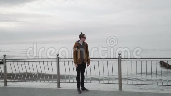 一个时髦的年轻人穿着棕色夹克戴着太阳镜微笑着看着大海视频的预览图