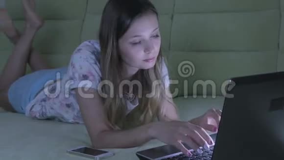 漂亮的女孩躺在沙发上用他的笔记本电脑工作她在智能手机上接电话视频的预览图