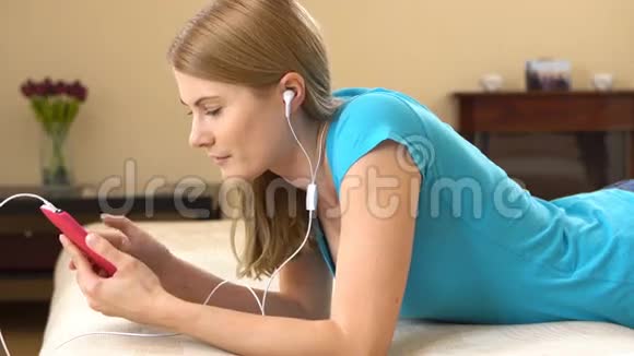 美丽迷人的年轻女人智能手机躺在沙发上听音乐视频的预览图