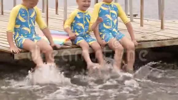 穿着泳衣的有趣的孩子坐在河边的木桥上摆动他们的腿创造喷雾男孩在度假时很开心视频的预览图