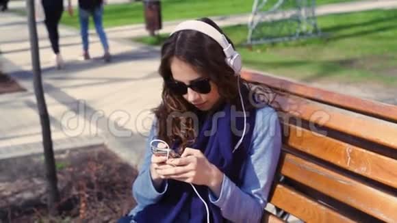 戴墨镜的年轻女子坐在长凳上用耳机听音乐视频的预览图