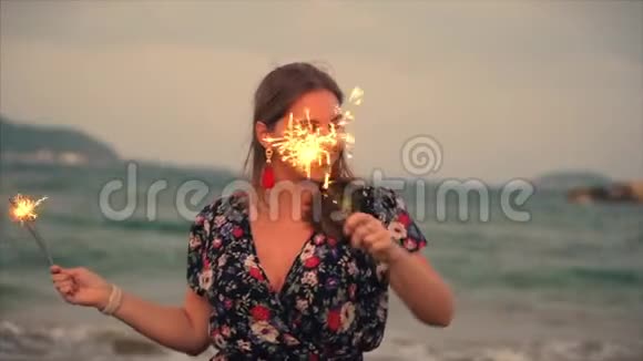 年轻快乐的微笑女人在日落时与火花在缓慢的运动中跳舞在海滩上的日落时与烟花一起跳舞视频的预览图