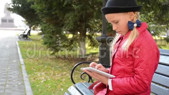 秋天在城市公园里带着平板电脑的小女孩坐在长凳上女孩在他的平板电脑上玩游戏视频的预览图