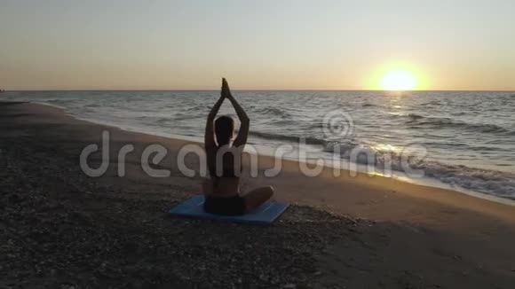 美丽的年轻女子坐在海滩上做瑜伽一边看日落视频的预览图