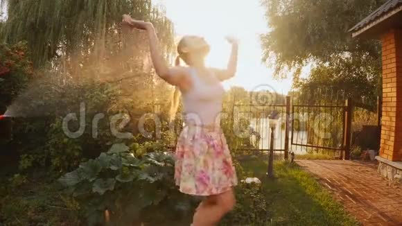日落时分年轻的笑女在花园水管下跳舞的慢镜头视频的预览图