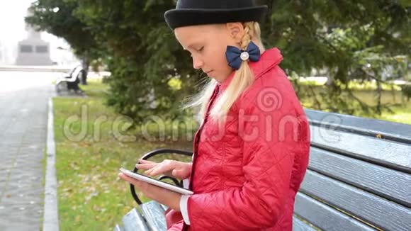 秋天在城市公园里带着平板电脑的小女孩坐在长凳上女孩在他的平板电脑上玩游戏视频的预览图
