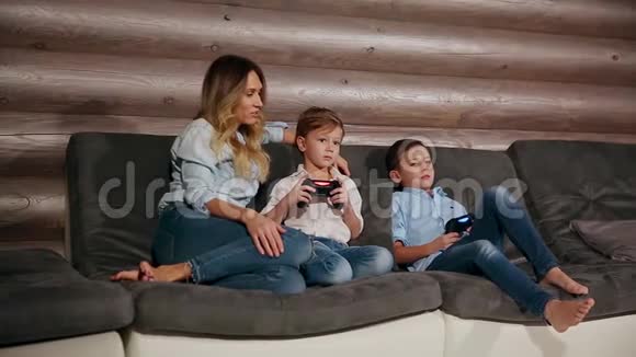 母亲和两个儿子坐在家里的沙发上用无线操纵杆玩电子游戏快乐的人们视频的预览图