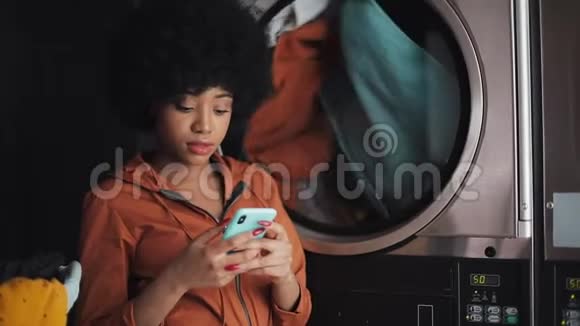在自助洗衣店用智能手机微笑的年轻女子视频的预览图