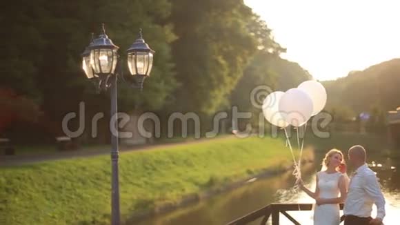 温柔的幸福和爱的时刻年轻漂亮的新婚夫妇带着气球拥抱在夕阳河桥上视频的预览图