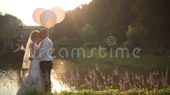 温柔的幸福时刻年轻漂亮的新婚夫妇带着气球拥抱在夕阳下的河底视频的预览图