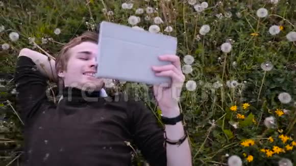 年轻快乐的学生在公园里用平板电脑躺在草地上的蒲公英微笑着笑视频的预览图