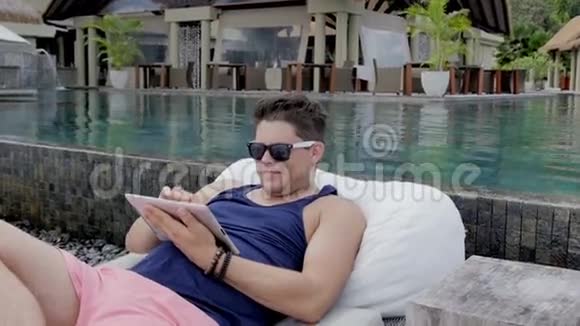 带墨镜的人坐在游泳池旁在热带度假胜地带数字平板电脑视频的预览图