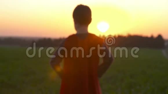 日落时穿着超级英雄斗篷的男孩站在田野里视频的预览图