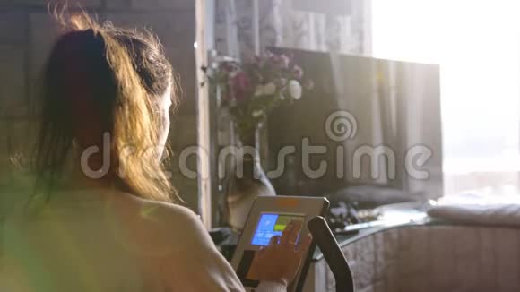 在日落时分年轻的黑发女子在靠窗的自行车模拟器上练习用镜头耀斑看着城市视频的预览图