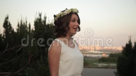 年轻漂亮的姑娘戴着花圈穿着白色连衣裙日落时分在户外唱歌跳舞视频的预览图