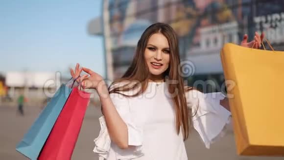 在现代玻璃建筑前穿着白色连衣裙的漂亮时尚模特摆着购物袋视频的预览图