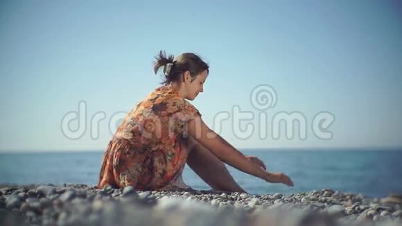 可爱的女孩坐在岸上把鹅卵石扔进海里视频的预览图