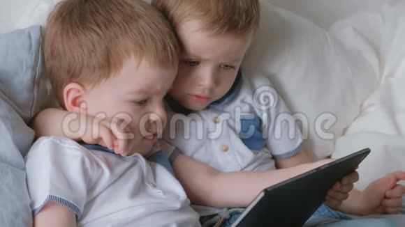 带平板电脑的孩子两个男孩双胞胎蹒跚学步看着躺在床上的平板电脑视频的预览图